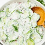 A white bowl filled with creamy cucumber salad, topped with fresh dill and small red onion pieces. A wooden spoon rests inside the bowl. The cucumber slices are coated in a creamy dressing.