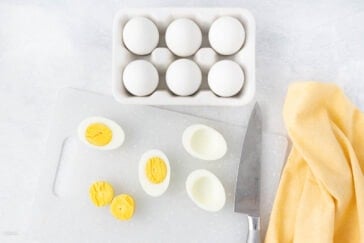 A carton of six white eggs is placed on a light surface. Next to it, a cutting board holds halved hard-boiled eggs, showing their yellow yolks. A knife lies nearby, and a yellow cloth is draped beside the board.
