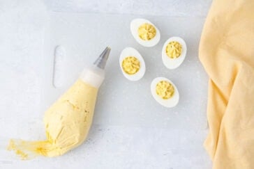 Piping bag filled with creamy yolk mixture next to four deviled eggs on a cutting board, with a yellow cloth nearby.