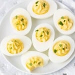 A plate of deviled eggs garnished with chopped parsley. One egg has a bite taken out of it, revealing a creamy yolk filling. The eggs are neatly arranged on a decorative white plate.