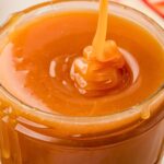 Close-up of caramel sauce being poured into a glass jar. The smooth, rich caramel forms swirls as it flows, with some drips running down the side of the jar, creating a glossy and appetizing texture.
