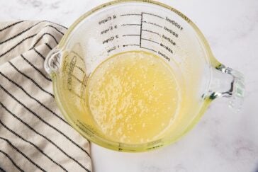 A glass measuring cup filled with a light yellow liquid sits on a marble surface. Next to it, a black and white striped cloth is partially visible.