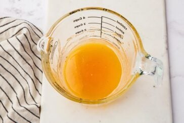A glass measuring cup filled with orange liquid sits on a white countertop. A striped cloth is partially visible on the left. The liquid is measured in cups and milliliters, visible through the transparent sides of the cup.