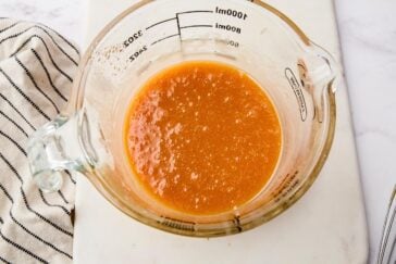 A glass measuring cup filled with a light brown, frothy liquid sits on a white surface, next to a striped cloth. The measuring cup has volume markings visible on its side.
