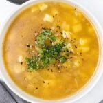 A bowl of soup with a clear broth, diced potatoes, and herbs. Garnished with fresh cilantro and black pepper.