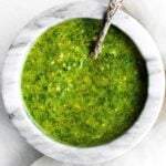 A marble bowl filled with vibrant green pesto sauce, displaying a slightly coarse texture. A decorative silver spoon rests in the pesto, adding an elegant touch to the presentation.