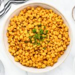 A white bowl filled with seasoned corn kernels, garnished with fresh cilantro. The dish is placed on a light surface with a striped cloth partially visible.