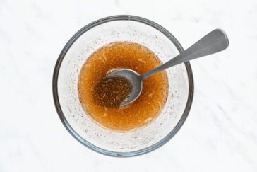 A glass bowl filled with a brown, speckled liquid sits on a white marble surface. A metal spoon rests inside the bowl, partially submerged in the liquid.