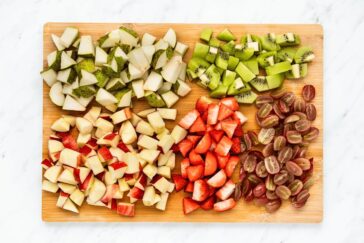 A wooden cutting board with diced fruit arranged in sections: pears, kiwis, strawberries, and grapes. The fruits are neatly organized and vibrant, resting on a white marble surface.