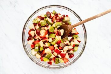 A glass bowl filled with a colorful fruit salad, featuring diced apples, pears, kiwi, strawberries, grapes, and pomegranate seeds. A wooden spoon is placed inside the bowl, all set on a marble surface.