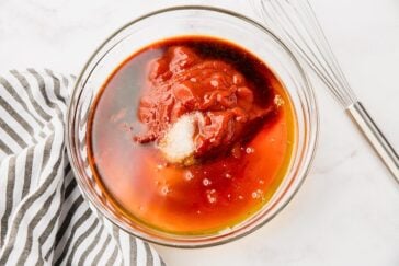 A clear glass bowl contains a mixture of ketchup, soy sauce, brown sugar, and possibly other liquid ingredients. It's placed on a white surface next to a striped kitchen towel and a whisk, indicating preparation for mixing.