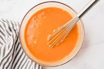 A bowl of creamy orange sauce with a wire whisk rests on a white marble surface. A black and white striped cloth is partially visible to the left.