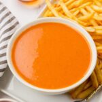 A white bowl filled with creamy, orange-colored fry sauce placed next to a serving of crispy, golden French fries on a white plate. A striped cloth is visible beside the bowl.