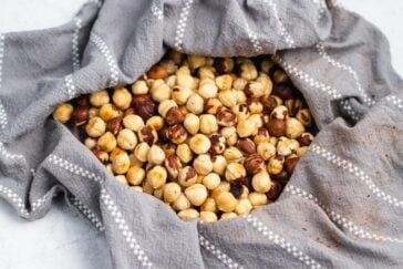 A pile of roasted hazelnuts wrapped in a gray cloth with white dotted lines. The cloth partially unfolds, revealing the mix of light and dark brown nuts on a smooth surface, showcasing how to roast hazelnuts perfectly.