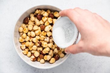 A hand demonstrating how to roast hazelnuts by carefully pouring a small bowl of sugar over a larger bowl of hazelnuts on a light countertop.