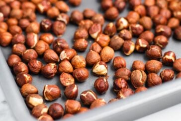 A baking tray filled with freshly roasted hazelnuts, demonstrating how to roast them perfectly. The nuts have a rich brown color, some with slightly cracked shells, highlighting their natural textures on the light countertop.