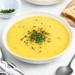 A bowl of creamy soup garnished with chopped herbs and ground pepper. A slice of bread is on a wooden board nearby. A spoon and a small bowl with more herbs are beside the soup on a white surface.