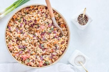 A large bowl of colorful macaroni salad with chopped vegetables and herbs, mixed with dressing. A wooden spoon is in the bowl. Nearby are bowls of peppercorns and salt with gold spoons. Green onions are in the background.