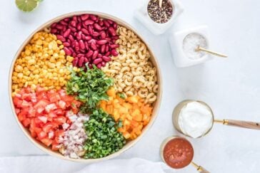 A bowl filled with corn, kidney beans, macaroni, chopped tomatoes, onions, bell peppers, and cilantro. Surrounding it are small containers with sour cream, salsa, salt, and peppercorns on a light surface.