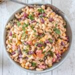 A bowl of creamy pasta salad featuring elbow macaroni, red kidney beans, corn, chopped tomatoes, and fresh herbs garnished on top, with a fork resting on the bowl.