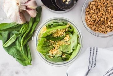 Ingredients for pesto are arranged on a white surface, including fresh basil leaves in a blender, garlic cloves, a small bowl of sunflower seeds, and a striped cloth napkin.