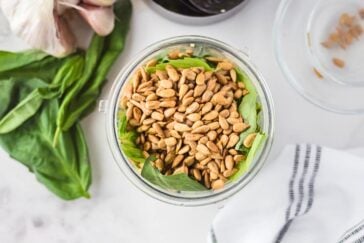 Top view of a food processor bowl filled with fresh basil leaves and pine nuts, placed on a white marble surface. Fresh garlic and basil leaves are on the side, with a white towel featuring black stripes nearby.