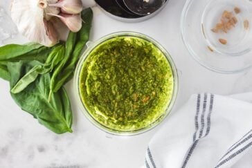 A small glass bowl of vibrant green pesto sits on a marble surface next to a bunch of fresh basil leaves, a garlic bulb, and a striped cloth. An empty bowl and black measuring cups are nearby.