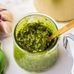 A jar of green pesto with a golden spoon inside, placed on a marble surface. A bulb of garlic and fresh basil leaves are nearby, along with part of a striped cloth. The pesto appears textured and fresh.