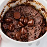 A close-up of a chocolate mug cake with melted chocolate chips on top, served in a white mug. The cake looks rich and moist, with a slightly textured surface.