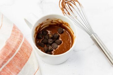 A white mug filled with chocolate cake batter and chocolate chips on top sits next to a whisk and a cloth with orange and white stripes on a marble surface.