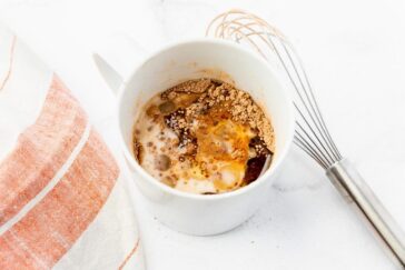 A white mug containing ingredients for a mug cake: milk, cocoa powder, and other dry ingredients, being mixed. A whisk and a red and white striped cloth are beside the mug on a white surface.