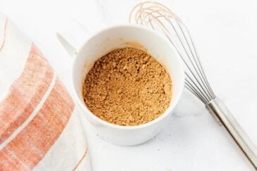 A white mug filled with brown sugar sits on a white surface next to a metal whisk. A white and orange striped cloth is partially visible on the left side.