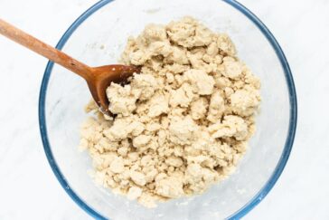 A glass bowl filled with crumbly dough mixture sits on a light surface. A wooden spoon is partially submerged in the dough, indicating mixing or preparation.