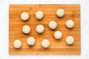 Twelve evenly spaced dough balls sit on a wooden cutting board, which is placed on a white marble surface. The dough balls are similar in size and color.