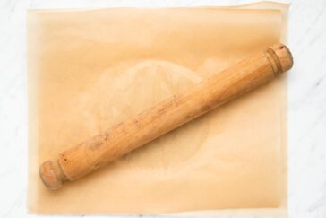 A wooden rolling pin rests diagonally on a sheet of parchment paper placed on a white marble surface.