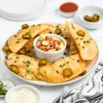 A plate of crispy, golden empanadas garnished with sliced jalapeños and chopped herbs surrounds a small dish of colorful salsa. Additional bowls of sauces and a striped towel are nearby on a white surface.