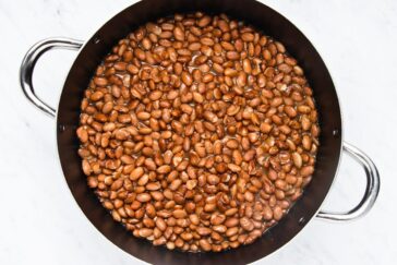 A large pot filled with cooked brown beans, submerged in their cooking liquid. The pot has shiny metal handles and is placed on a light surface.