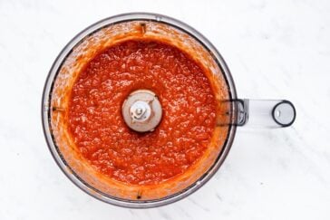 A top view of a food processor containing freshly blended tomato sauce. The sauce is bright red and evenly textured, surrounding the central blade. The processor is set on a light marble countertop.