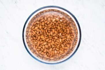 A bowl filled with pinto beans soaking in water on a white marble surface.