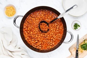 A pot of baked beans with a ladle on a marble countertop. Surrounding the pot are a bowl of grated cheese, a plate, chopped herbs on a small dish, a knife, and a wooden board with herbs. A cloth is placed nearby.