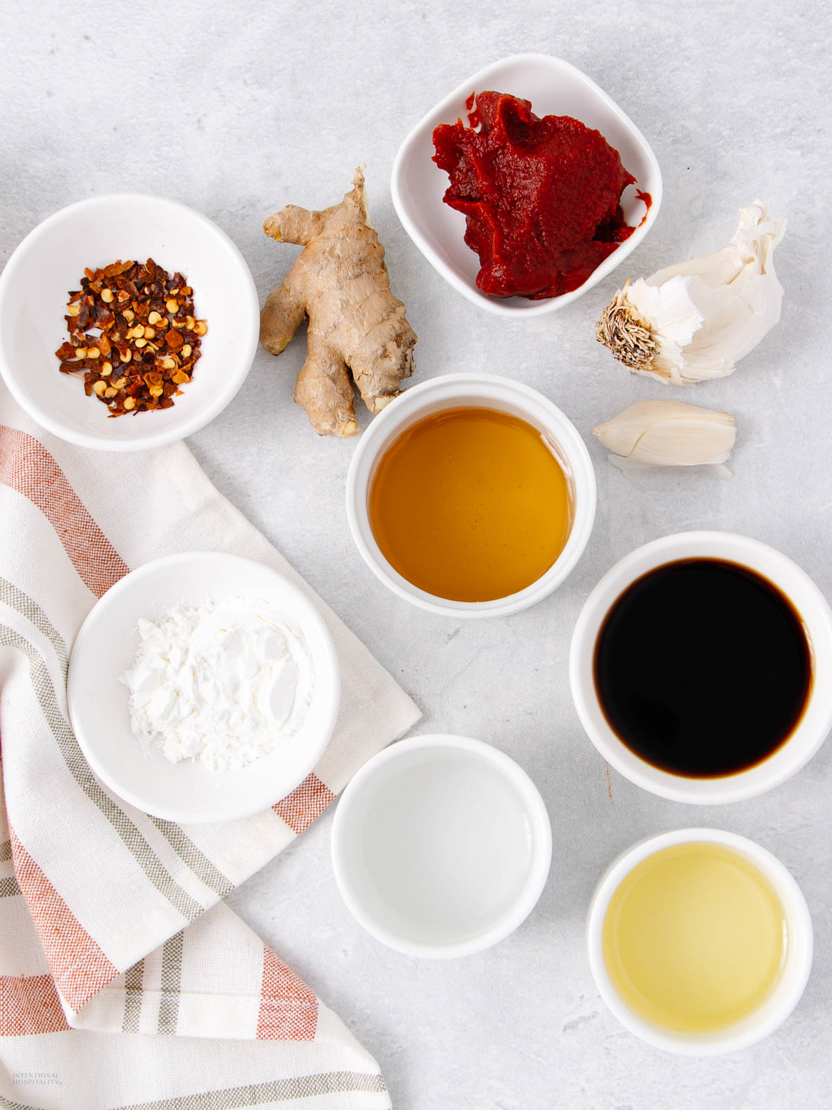 Various sauce ingredients in small bowls, perfect for creating a homemade stir fry sauce—soy sauce, honey, vinegar, red chili flakes, cornstarch, tomato paste, ginger root, and garlic—are arranged on a light surface with a striped cloth.