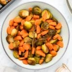 A bowl of roasted Brussels sprouts and sliced carrots garnished with sprigs of rosemary, placed on a marble surface. A roasted garlic bulb is partially visible in the corner.
