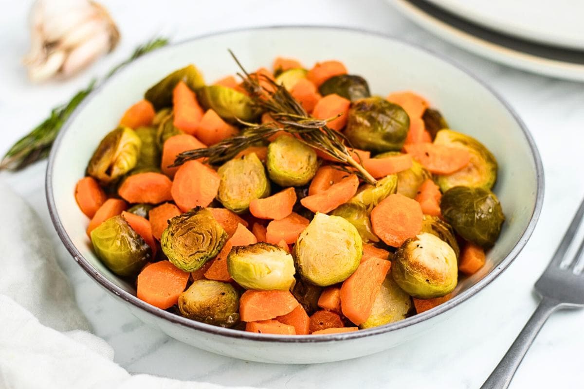 A bowl of roasted Brussels sprouts and sliced carrots, garnished with rosemary sprigs, on a white marble surface with a fork nearby.