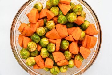 A glass bowl filled with chopped Brussels sprouts and sweet potatoes. The vegetables are seasoned and ready to be roasted.