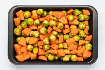 A baking tray filled with halved Brussels sprouts and cubed sweet potatoes, seasoned and ready for roasting.