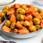 A bowl filled with roasted Brussels sprouts and sweet potato chunks, garnished with a sprinkle of seasoning. The dish is served with a large spoon, placed on a light-colored surface near a checkered cloth.