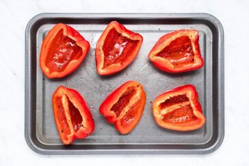 Halved red bell peppers arranged on a dark baking tray, seasoned with oil and spices, ready for roasting. The tray is placed on a light marble surface.