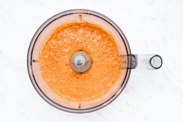 A top view of an orange sauce being blended in a transparent food processor on a white marble surface.