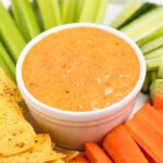 A white bowl filled with creamy orange dip, surrounded by fresh vegetables like celery, cucumber, and carrot sticks, along with tortilla chips on a platter.