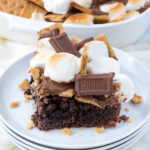 A slice of s'mores cake on a white plate, topped with toasted marshmallows, graham cracker pieces, and chocolate squares. Crumbs are scattered around the plate.